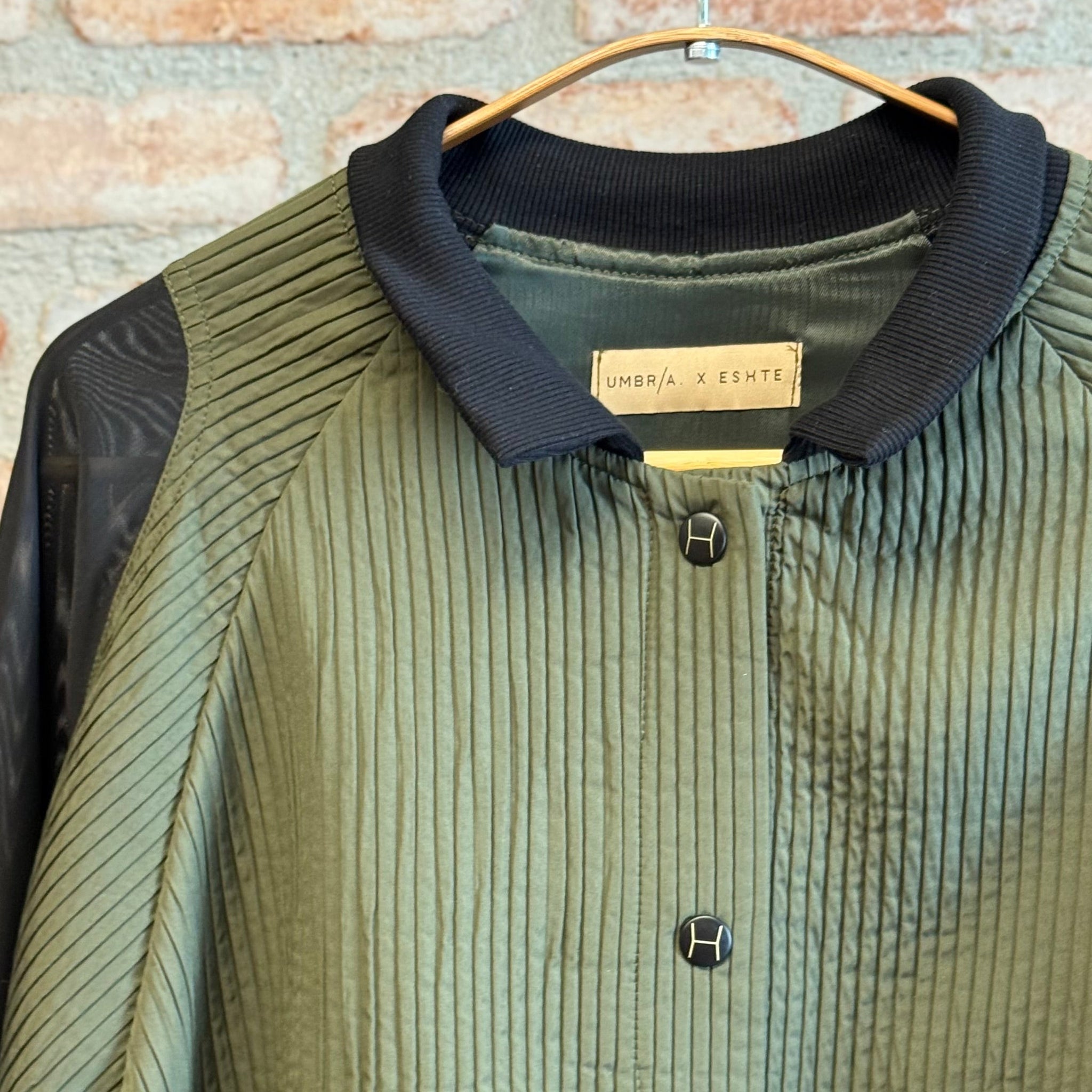 Green corduroy jacket with black collar on a hanger against a brick wall.